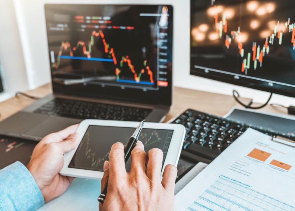 A close up of person's hands analyzing financial data on a tablet, with multiple monitors displaying stock market charts in the background.
