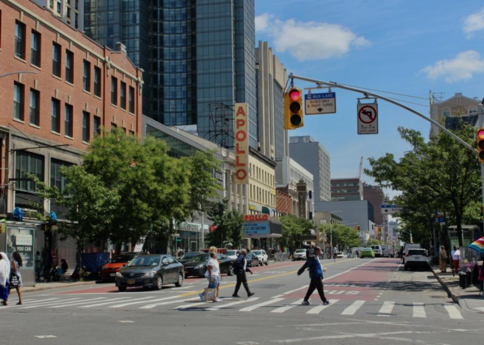 A city street with a mix of historic and modern buildings. The prominent "APOLLO" sign on one of the buildings. The street is active with pedestrians crossing the road, vehicles in motion, and various storefronts lining the sidewalk. 