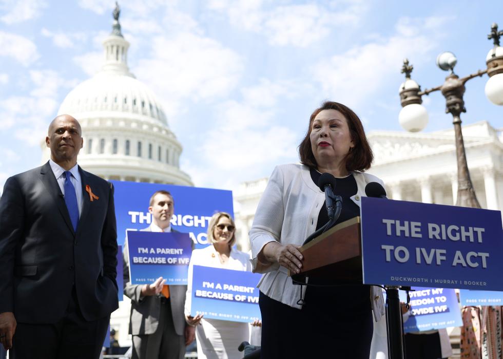 Senator Tammy Duckworth speaks alongside Senator Cory Booker during a news conference on access to IVF treatments outside of the U.S. Capitol Building. The podium in front of her has a poster that reads, The Right to IVF Act while people behind her hold signs that say, I'm a parent because of IVF.