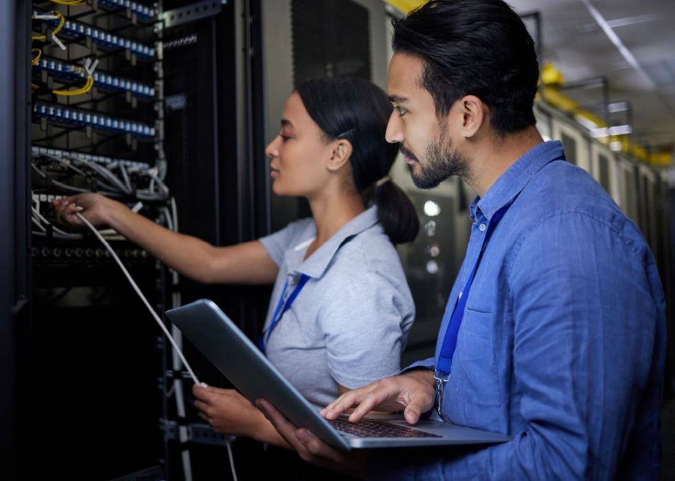 A young woman, holding a white cable, examines the IT equipment while a man beside her holds a laptop in a server room.