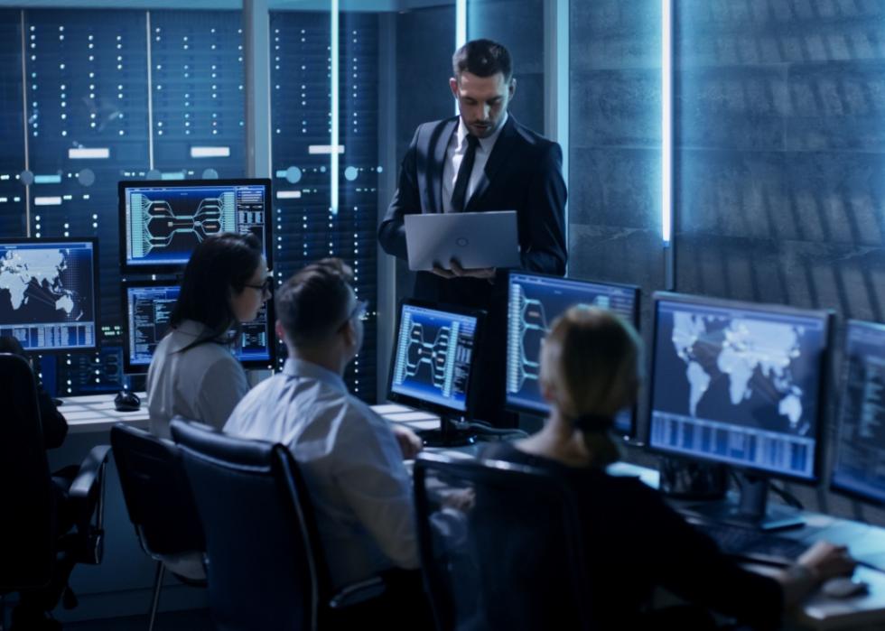 Three people sitting at desks in a dark lit room looking at the multiple screens in front of them. One man holding a laptop is facing three other people in the room.