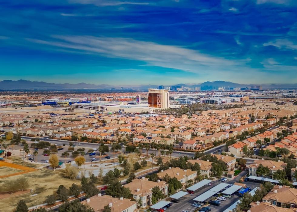 An aerial view of a sprawling suburban landscape in a sunny climate. The image features a vast expanse of residential homes with a variety of styles, including single-family houses and townhomes.