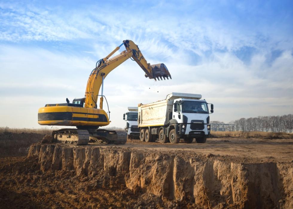 A huge yellow crawler excavator and a construction dump truck standing next to it while working on a sunny day against a blue sky.