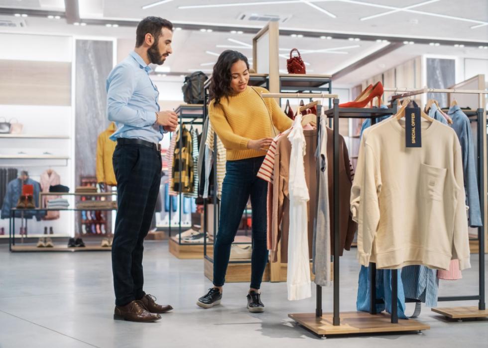 Young woman customer looking at pieces of clothing in a store, retail sales associate is standing next to her.