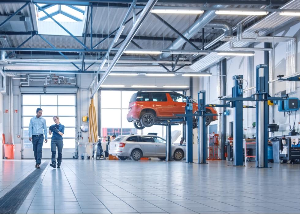 A woman and man walking in an industrial facility. One car is on the floor while the other one  is suspended high.