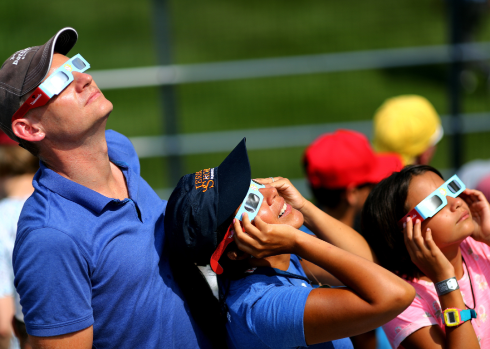 A man and two children are wearing paper glasses looking up into the sky. 