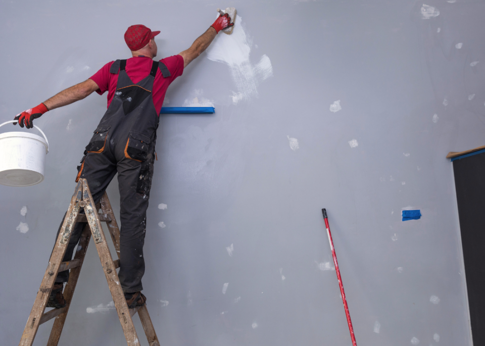A man in overalls and a hat stands on a ladder with a paint brush in his right hand and a bucket in his left.