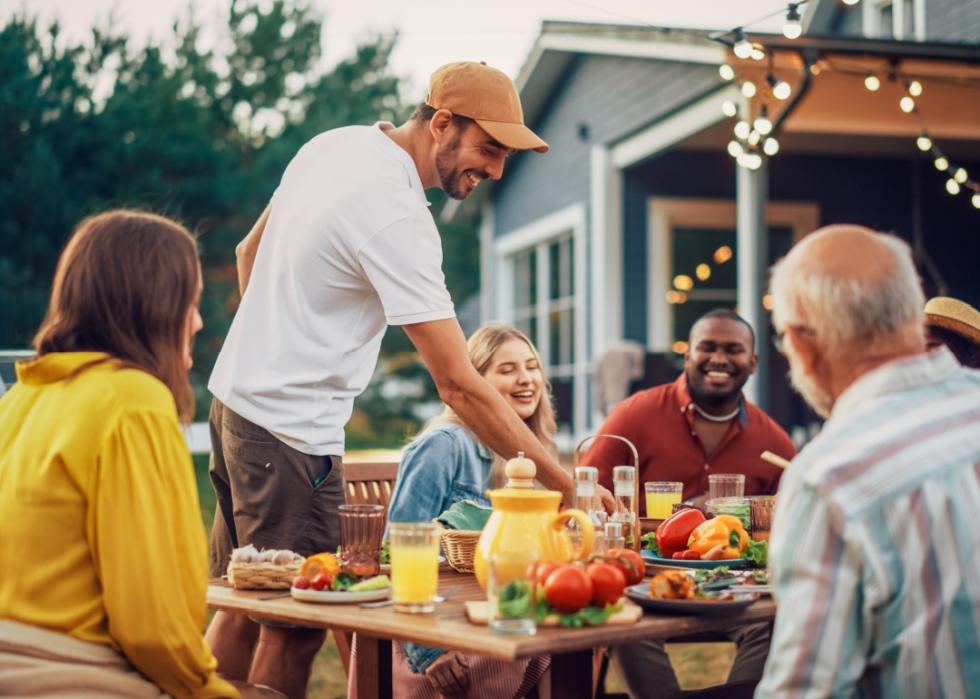 A young man smiling, leaning over the outdoor table. Group of people sitting around the table with drinks and food.