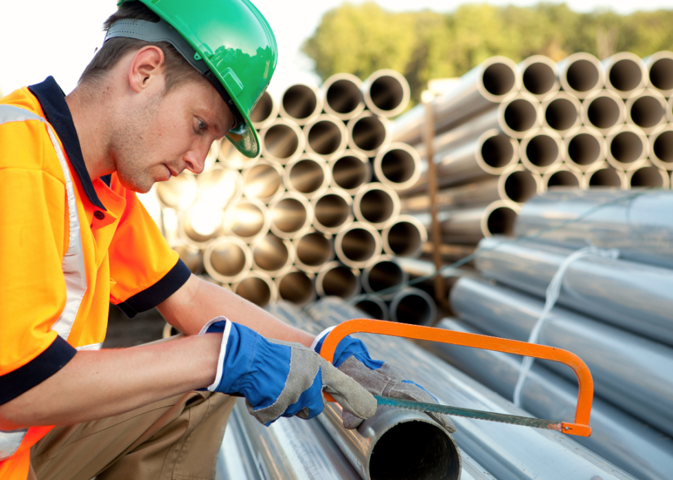 A man wearing a green hard hat cutting a long, metal pipe with a hand saw.
