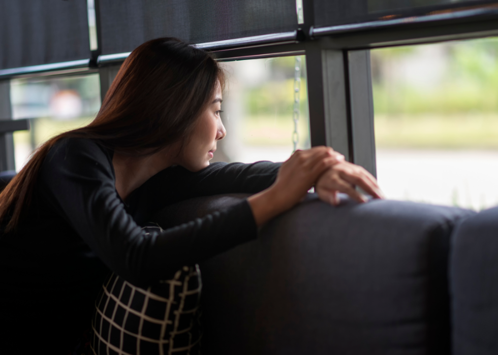 A bored woman sits on a couch peering out a window.