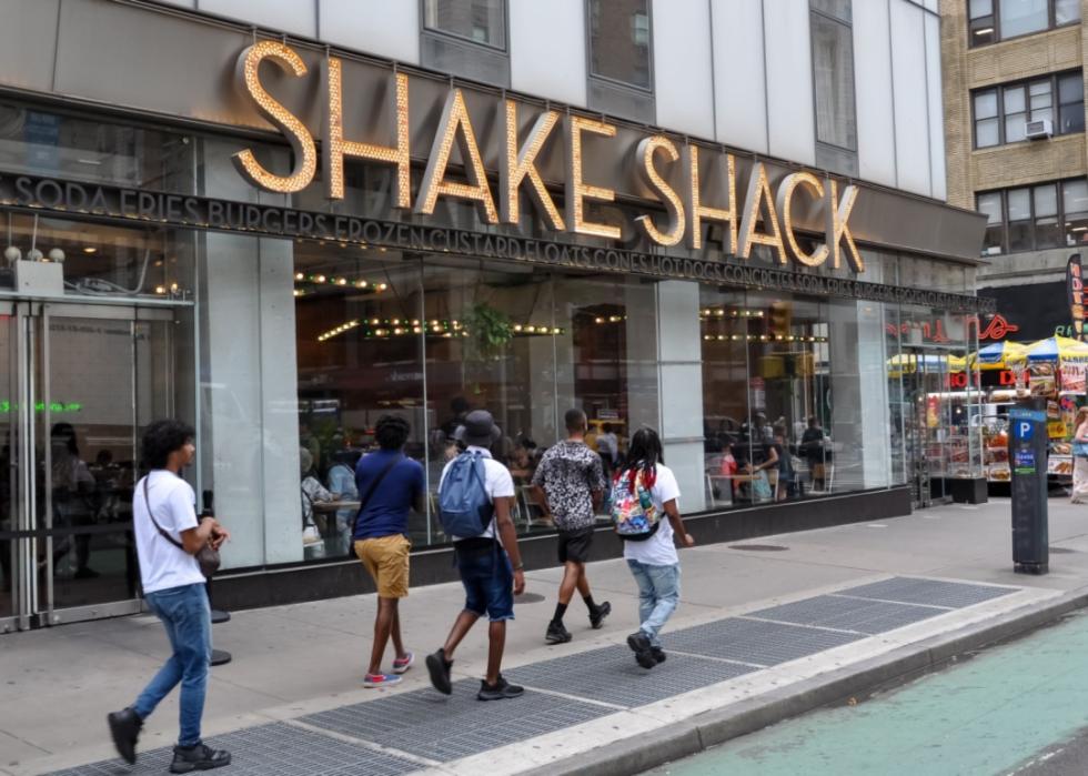 The exterior of a Shake Shack restaurant on an urban street with several people walking past it on the sidewalk.