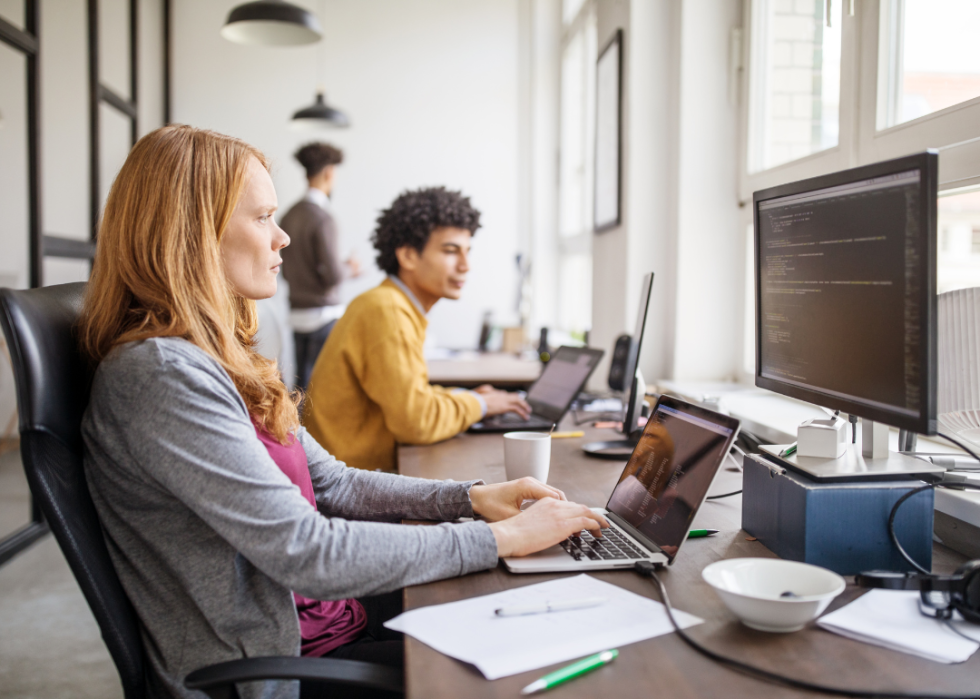 Two young software engineers are sitting at desks in a modern office, looking at large screens in front of them.