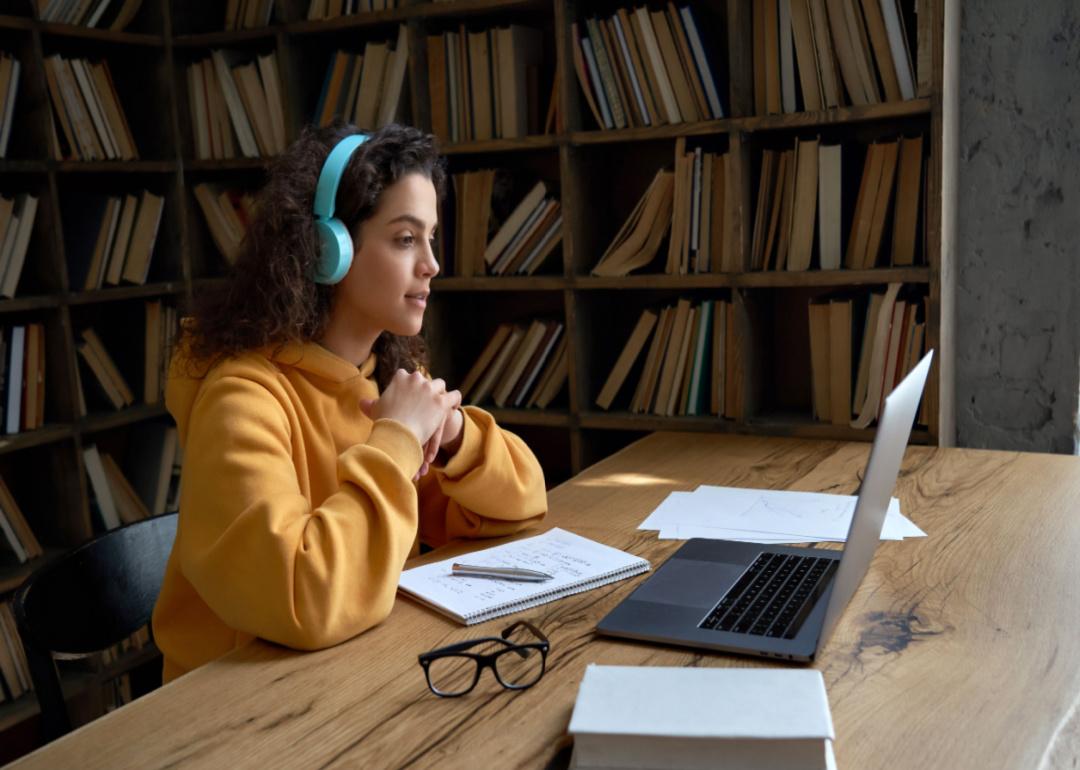 A college student wearing headphones and watching her laptop in a library.