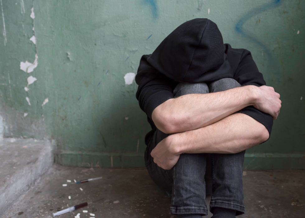 A teenager with their head resting on their knees sits on the ground against a deteriorating wall, with pills and syringes on the ground next to them.