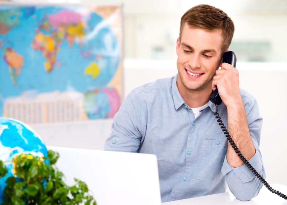 A young, smiling man sitting at the desk while holding a phone to his ear and looking at a laptop.
