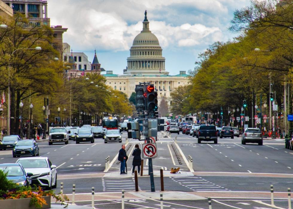 A large building with the white dome in center. The street in the foreground is busy with traffic, featuring multiple lanes filled with cars, and traffic lights are visible in the middle of the road. On either side, trees line the street.