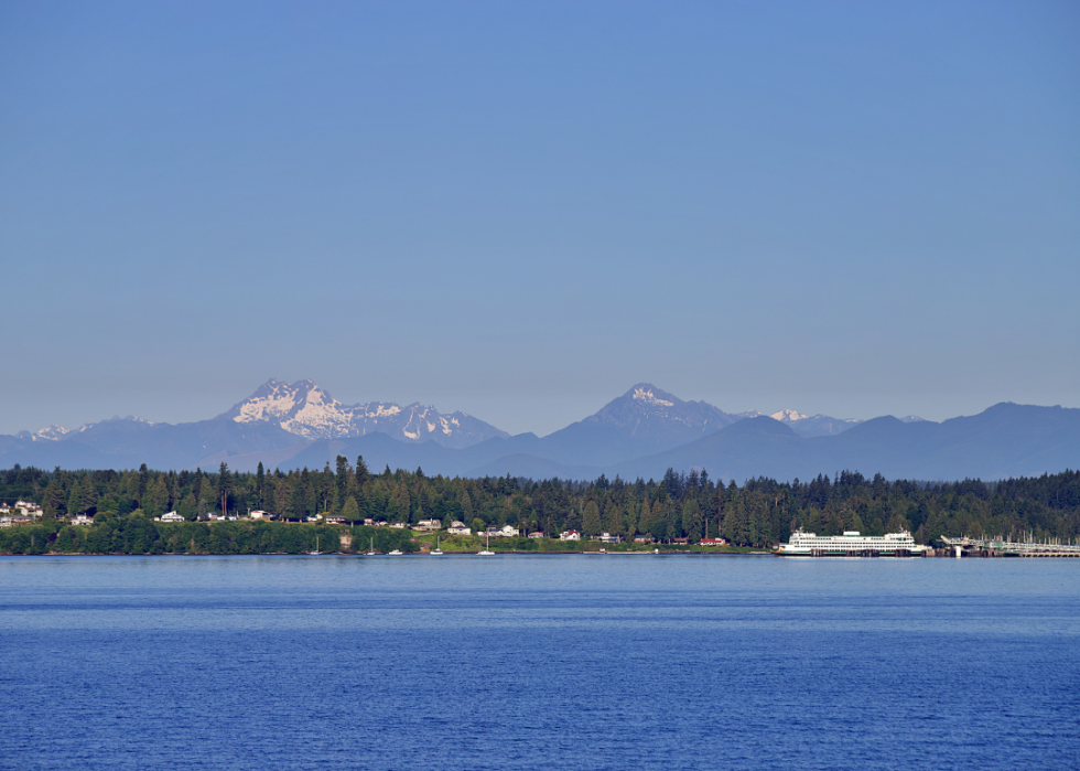 A town situated by the body of water with tall snowy mountain peaks and green trees in the background.