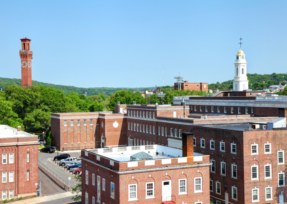 A high-up view of a town with red brick buildings and thick tree-covered land in the background.