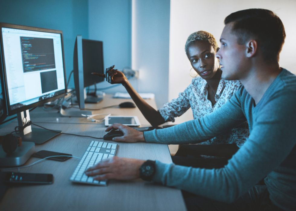 A man looking at the computer screen in a dimmed office; a woman sitting next to him looking in his direction while pointing at her computer screen.