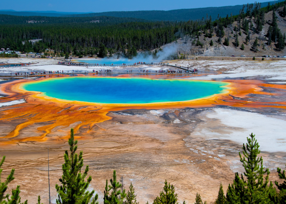 An aerial view of vibrant turquoise hot spring with outer ring in bright orange and green mountains in the background.