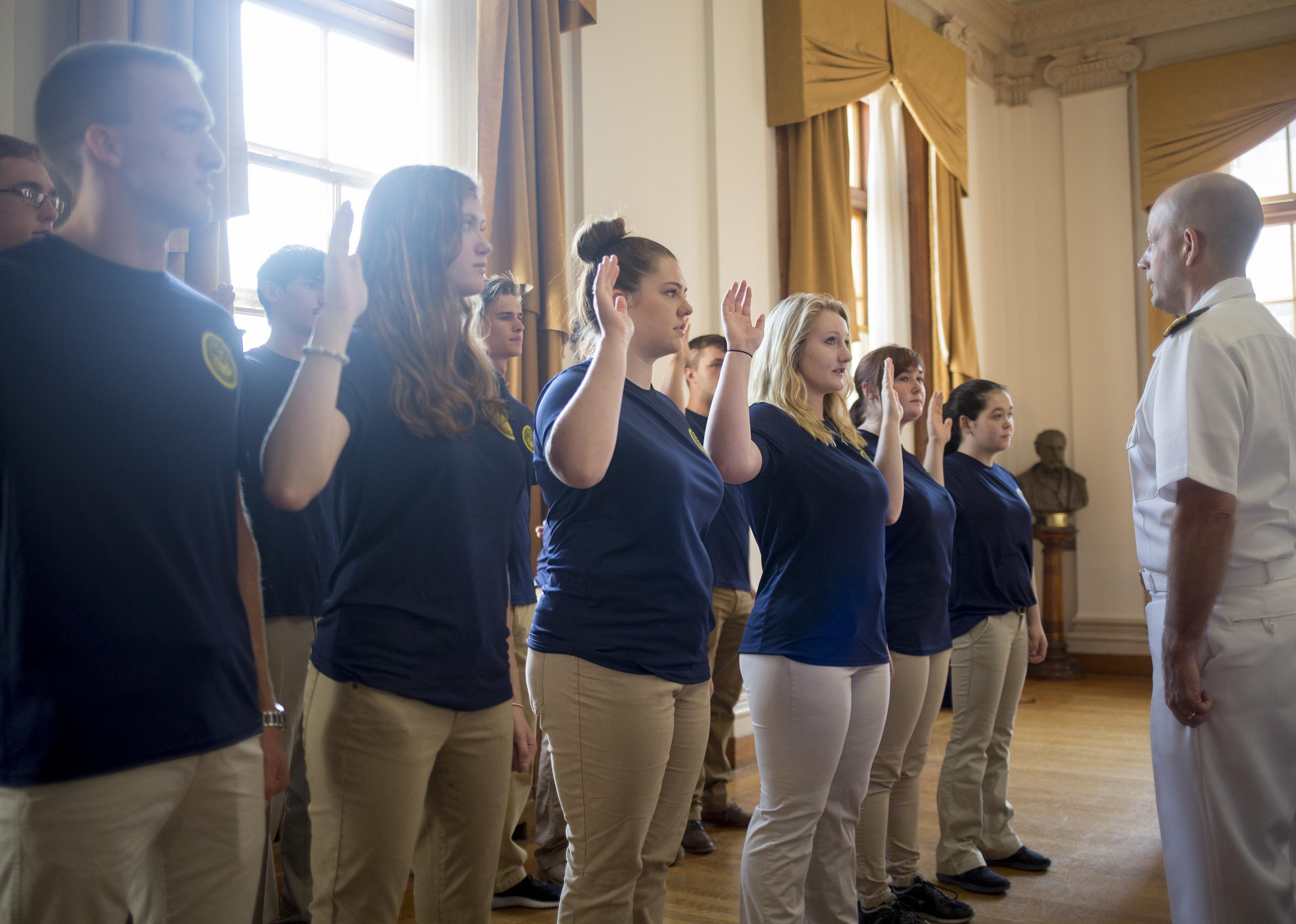 New Navy sailors in blue shirts and khaki pants are sworn in by a sailor in white.