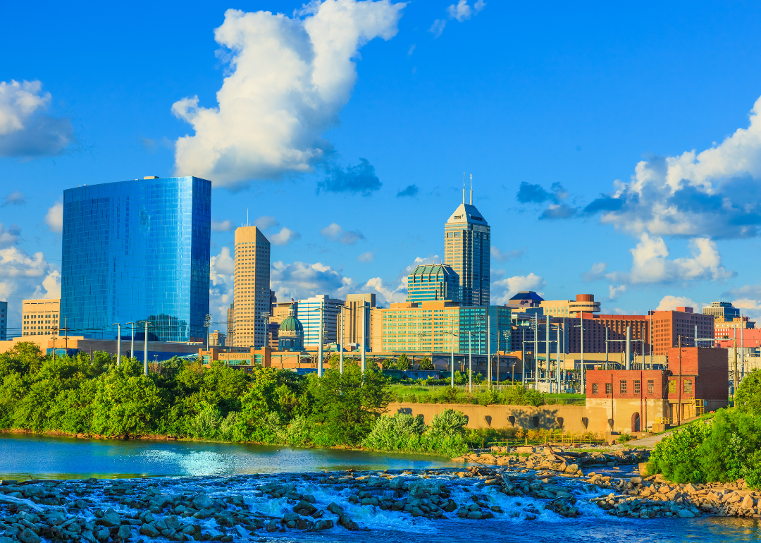 Indianapolis, IN skyline with blue sky behind and a rocky river in the foreground.