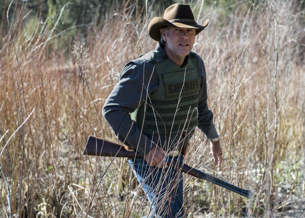 A man in a cowboy hat and bullet proof vest holding a long gun.