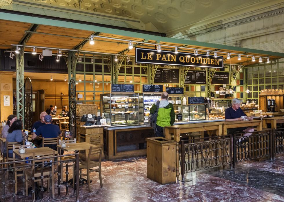 The interior of a Le Pain Quotidien bakery with a customer waiting to be served while other customers sit in. 