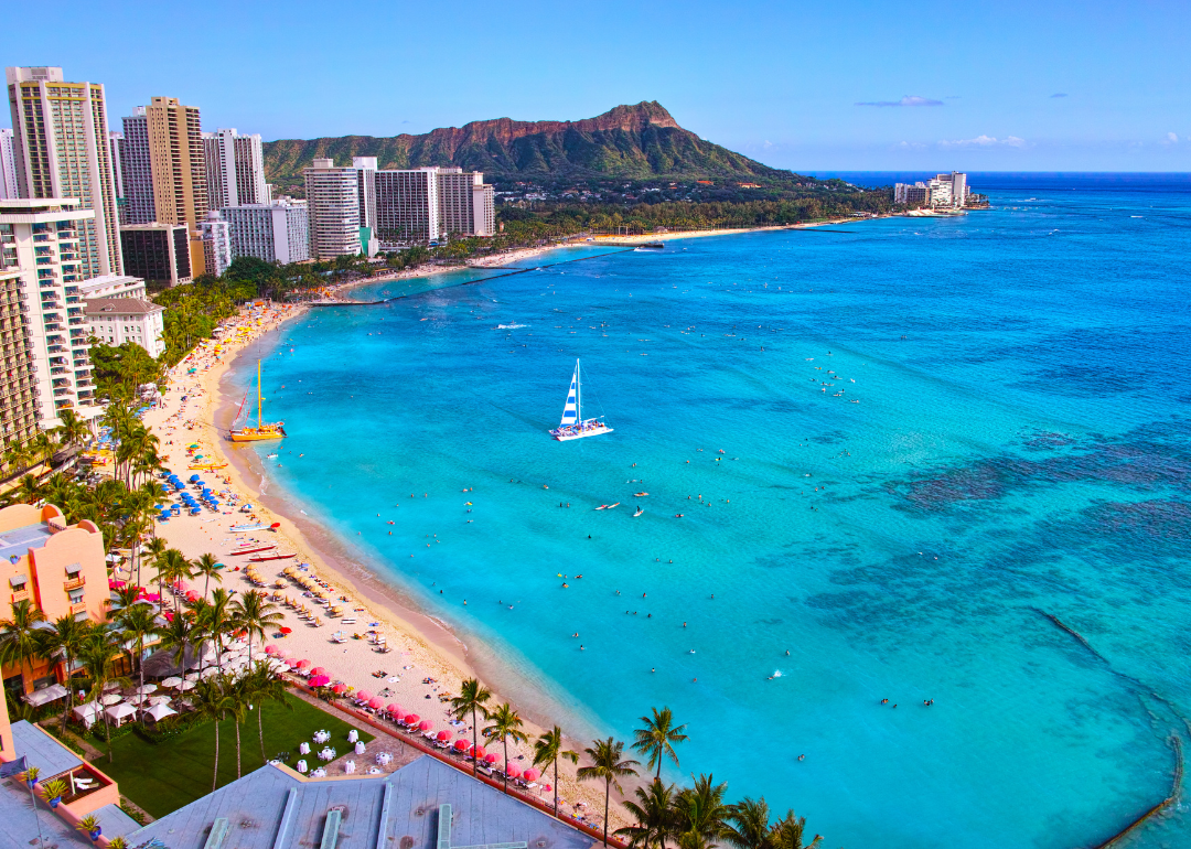 Boats on turquoise water and umbrellas on a crowded beach next to tall buildings.