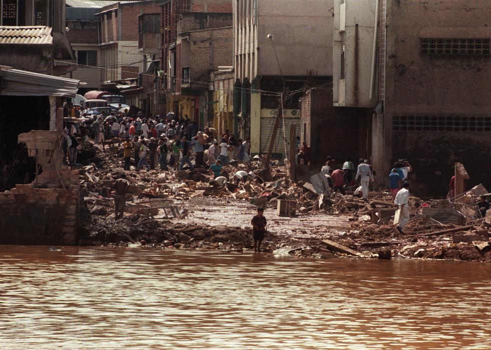 People gather in the streets of an urban area, surrounded by wreckage as the flood waters near.