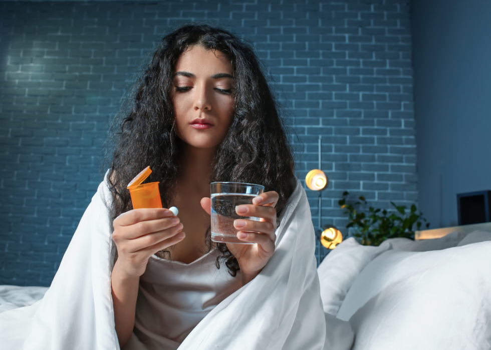 Woman on the edge of the bed looking at a prescription and holding a glass of water.