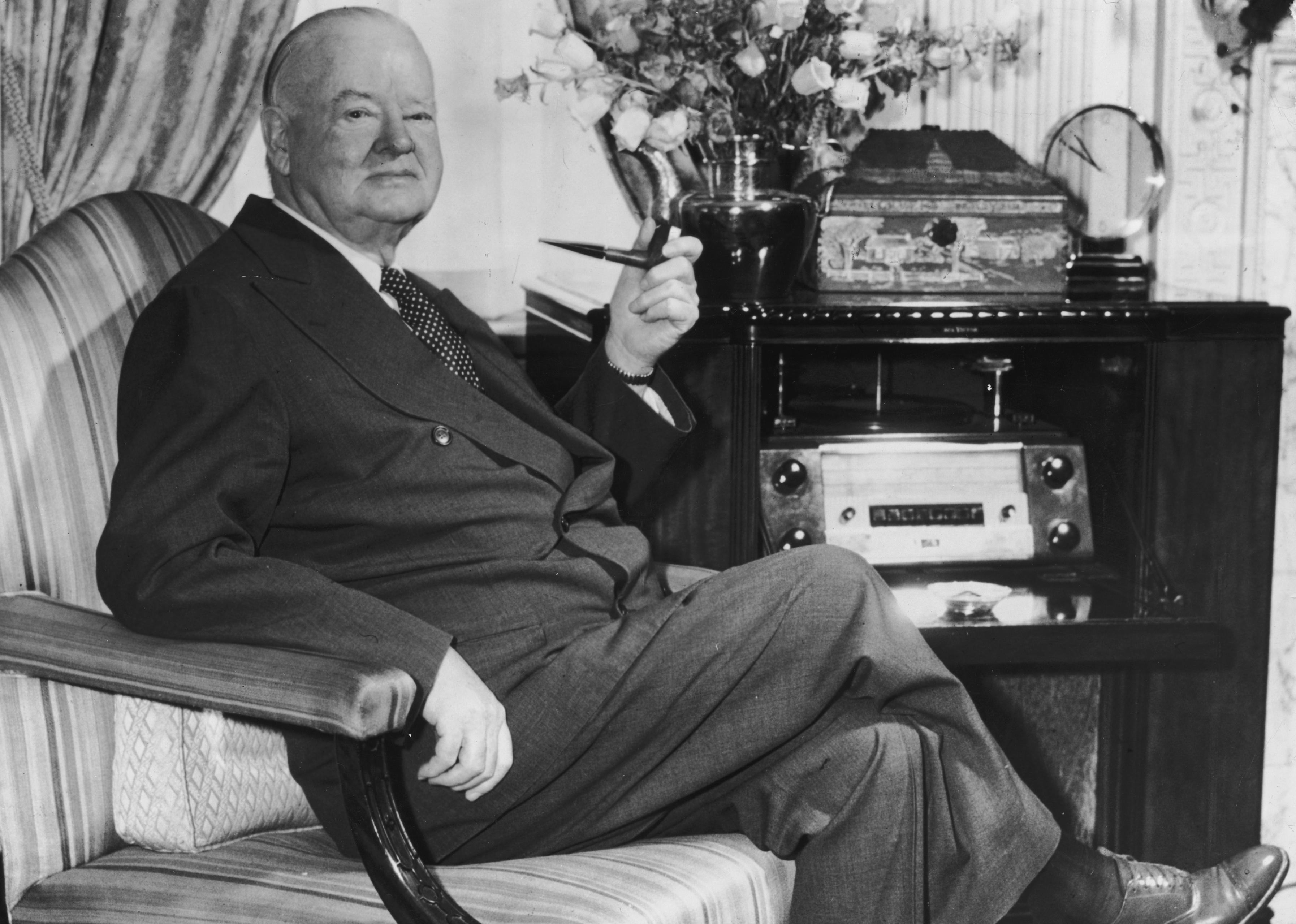 Herbert Hoover in a chair with legs crossed holding a pipe.