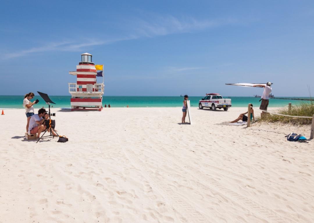 A photographer and their assistants taking photos of someone on the beach with a red and white striped lifeguard stand  by the water.