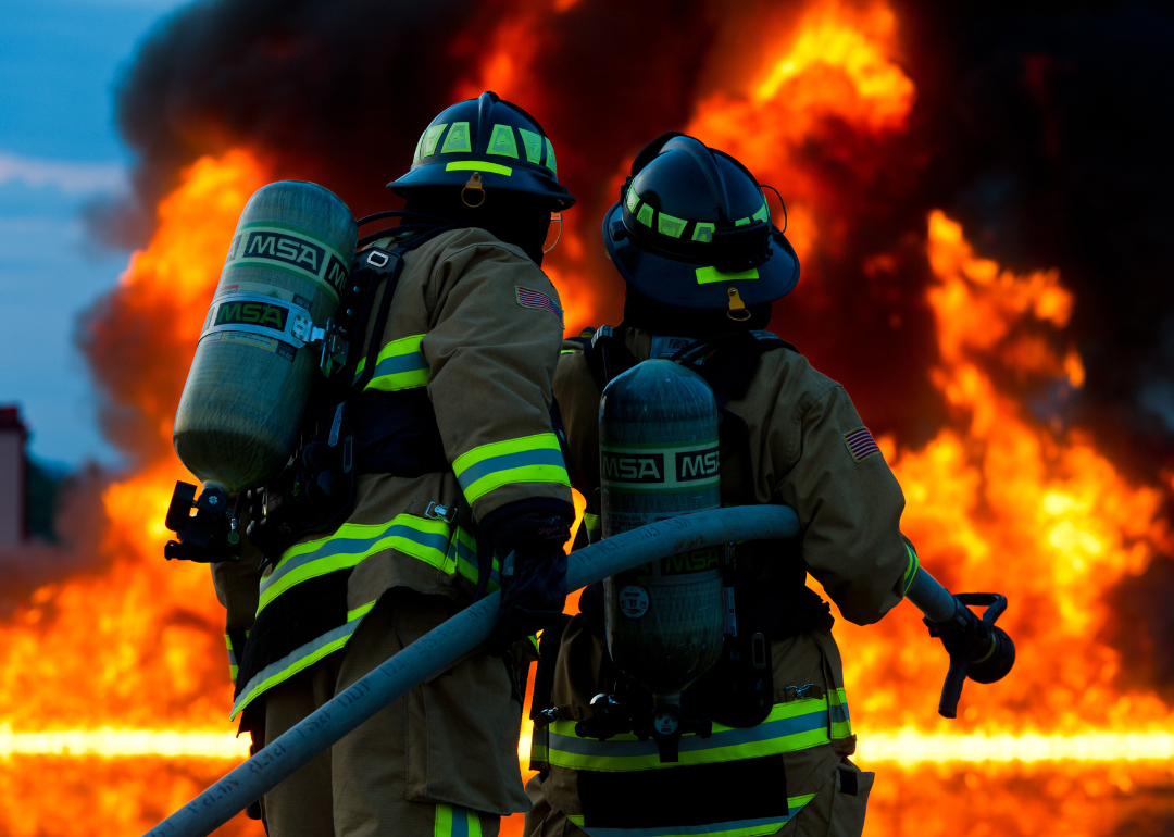 Two firefighters hold a hose in front of a huge flame.