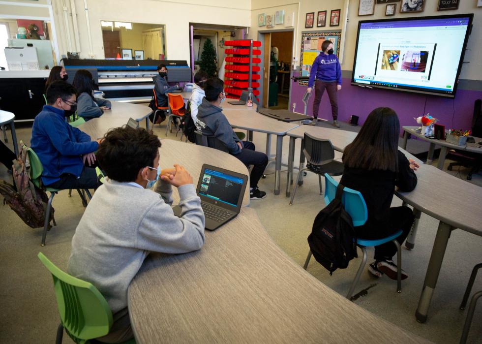 Students back in class and socially distanced at Olive Vista Middle School as the teacher shows slides on a large TV screen.