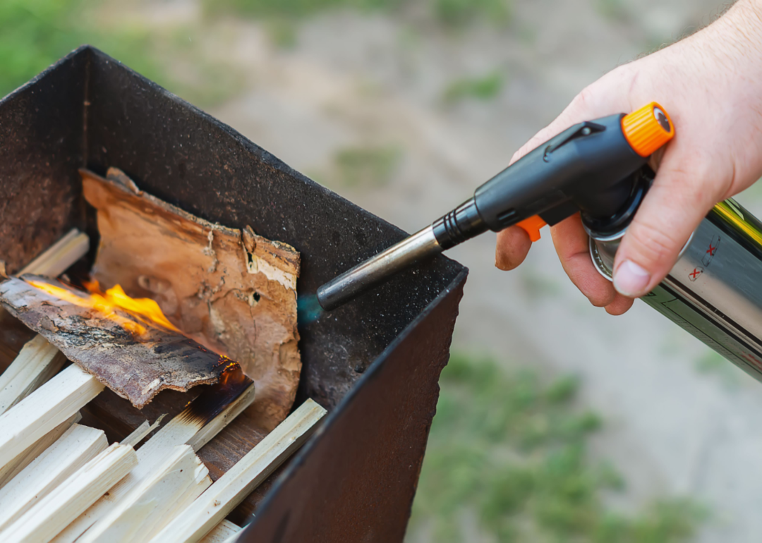 A hand holding a torch cylinder to light some sticks on a fire.