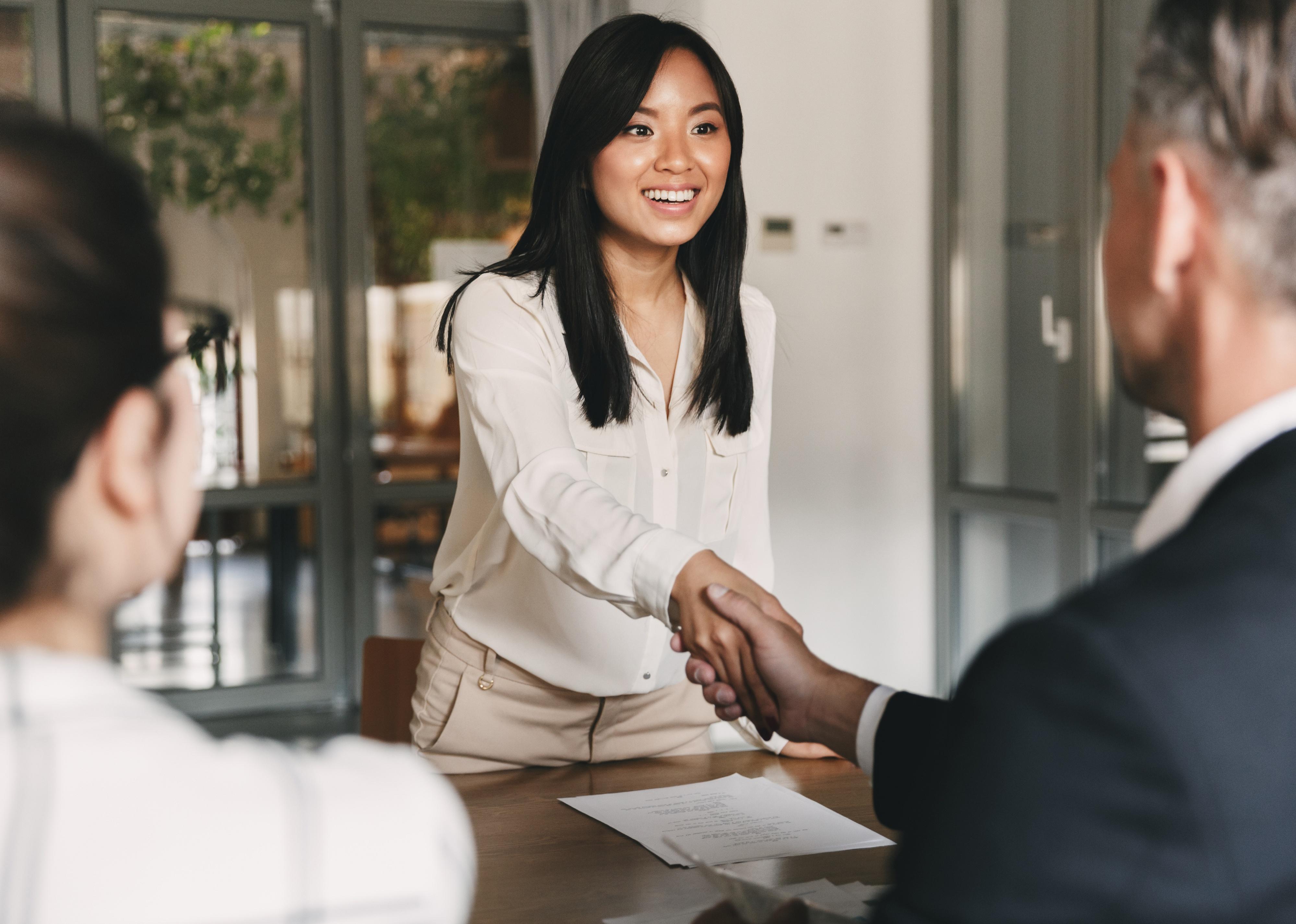 Back view of two employers sitting in an office and shaking a young woman's hand.