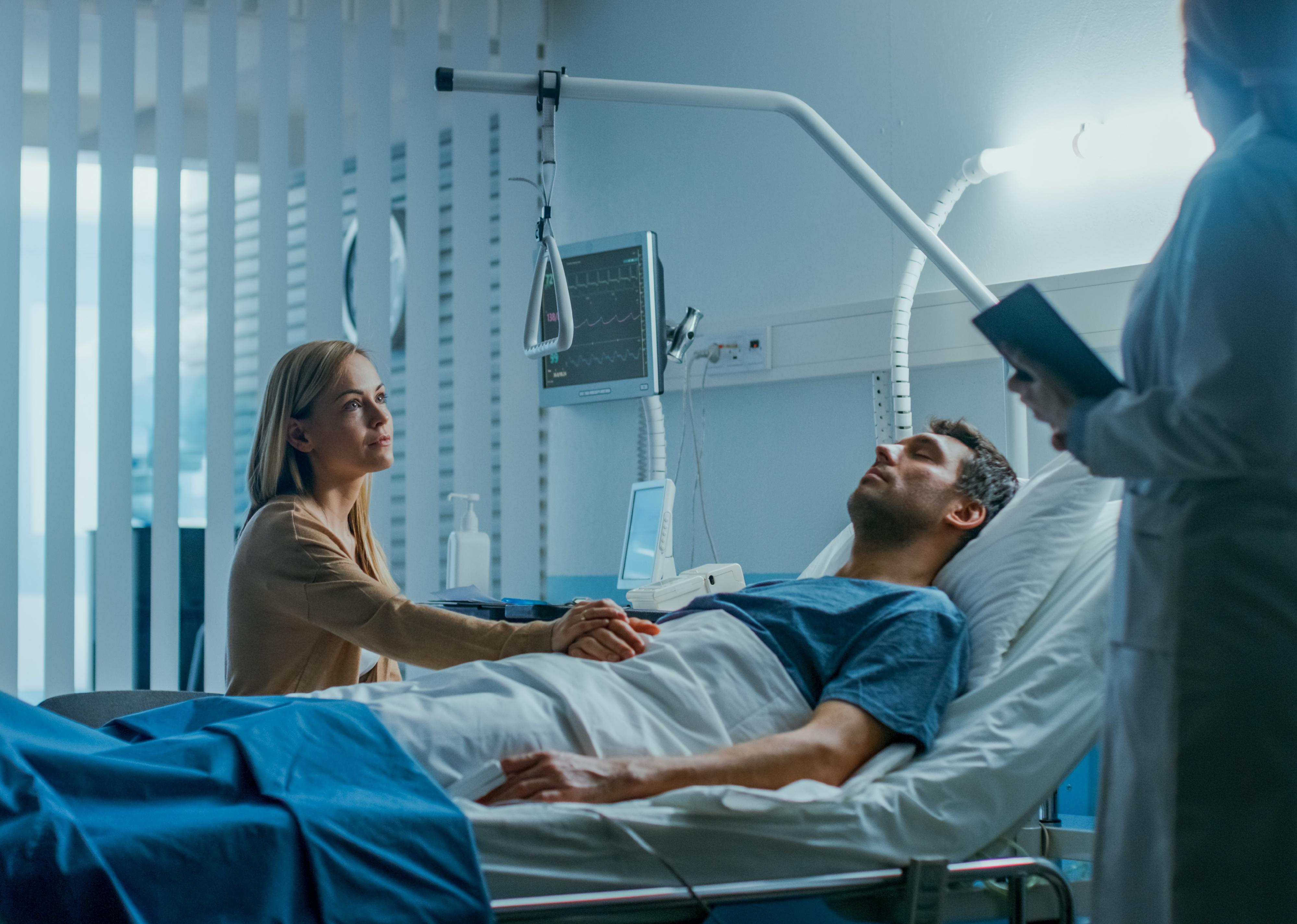 Patient lying in bed with a visitor sitting beside him while a doctor speaks.