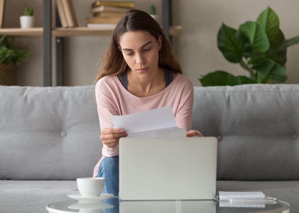 Woman sitting on couch and holding paper with open laptop 