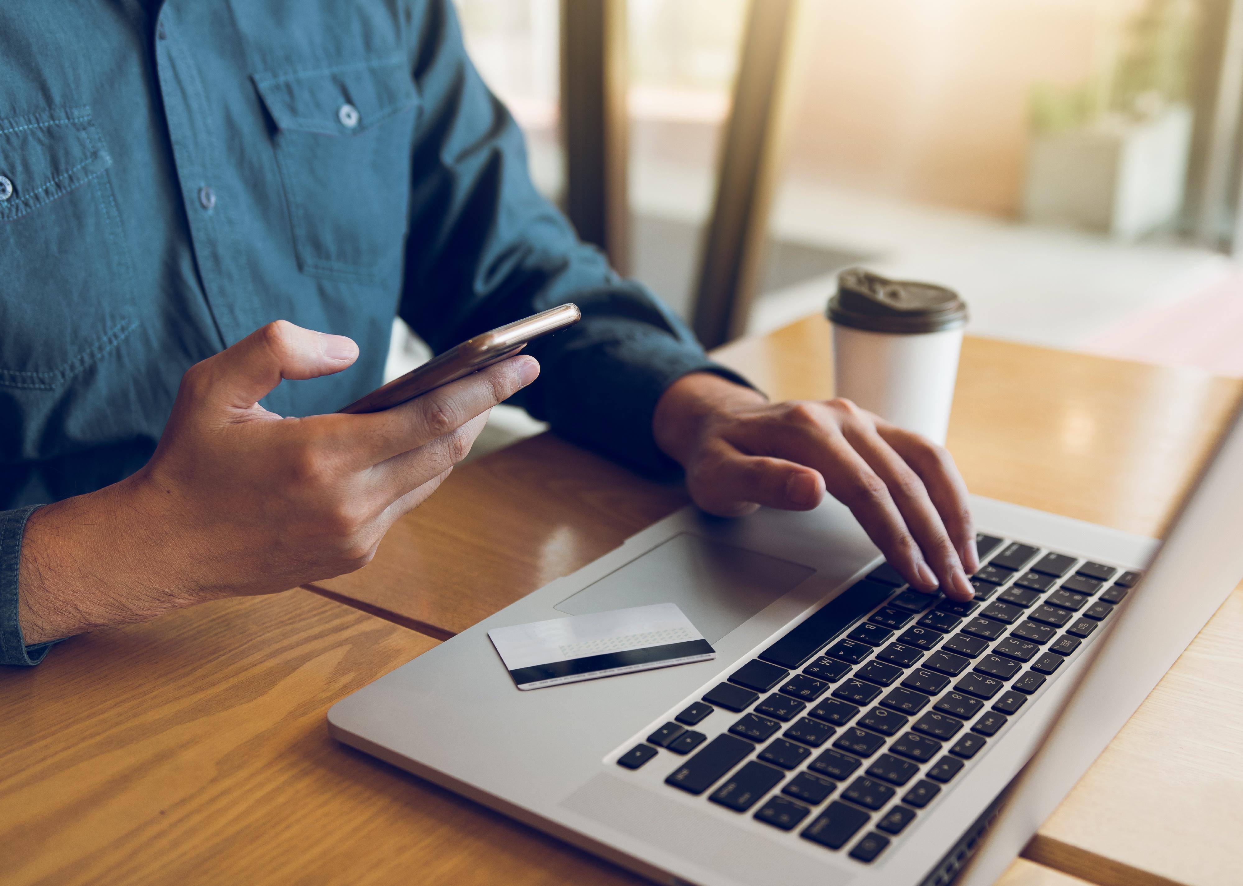 Man holding mobile phone while typing on laptop