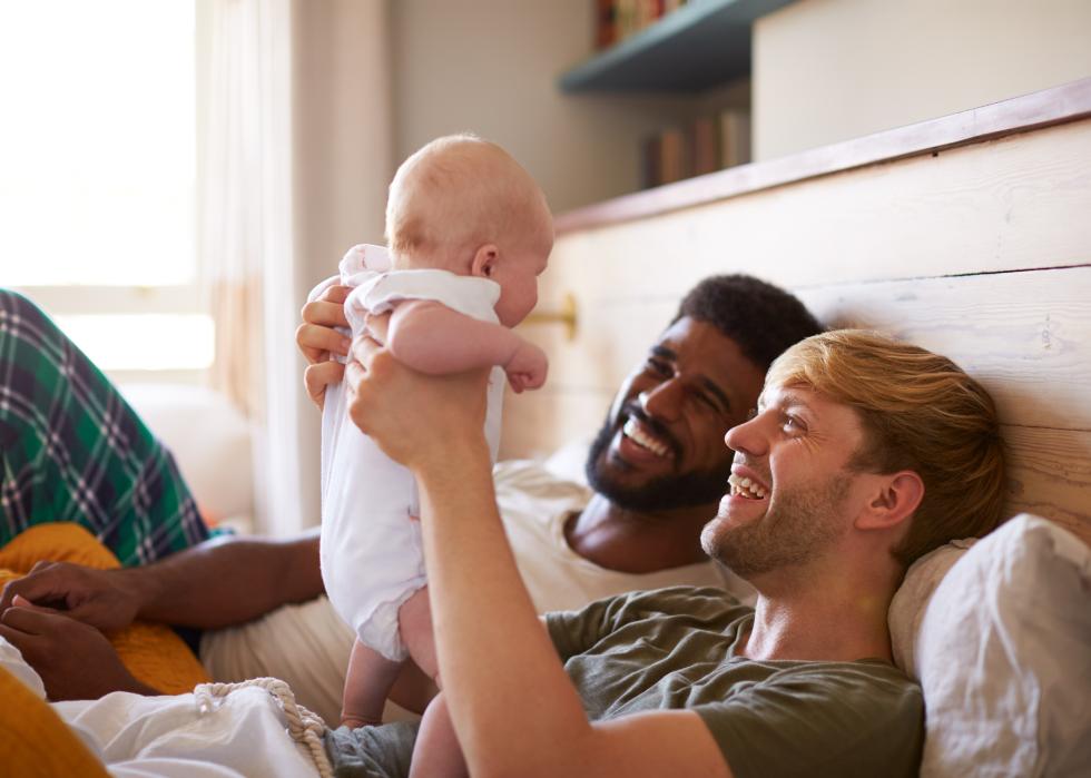Two smiling men laying in bed, one holding a baby