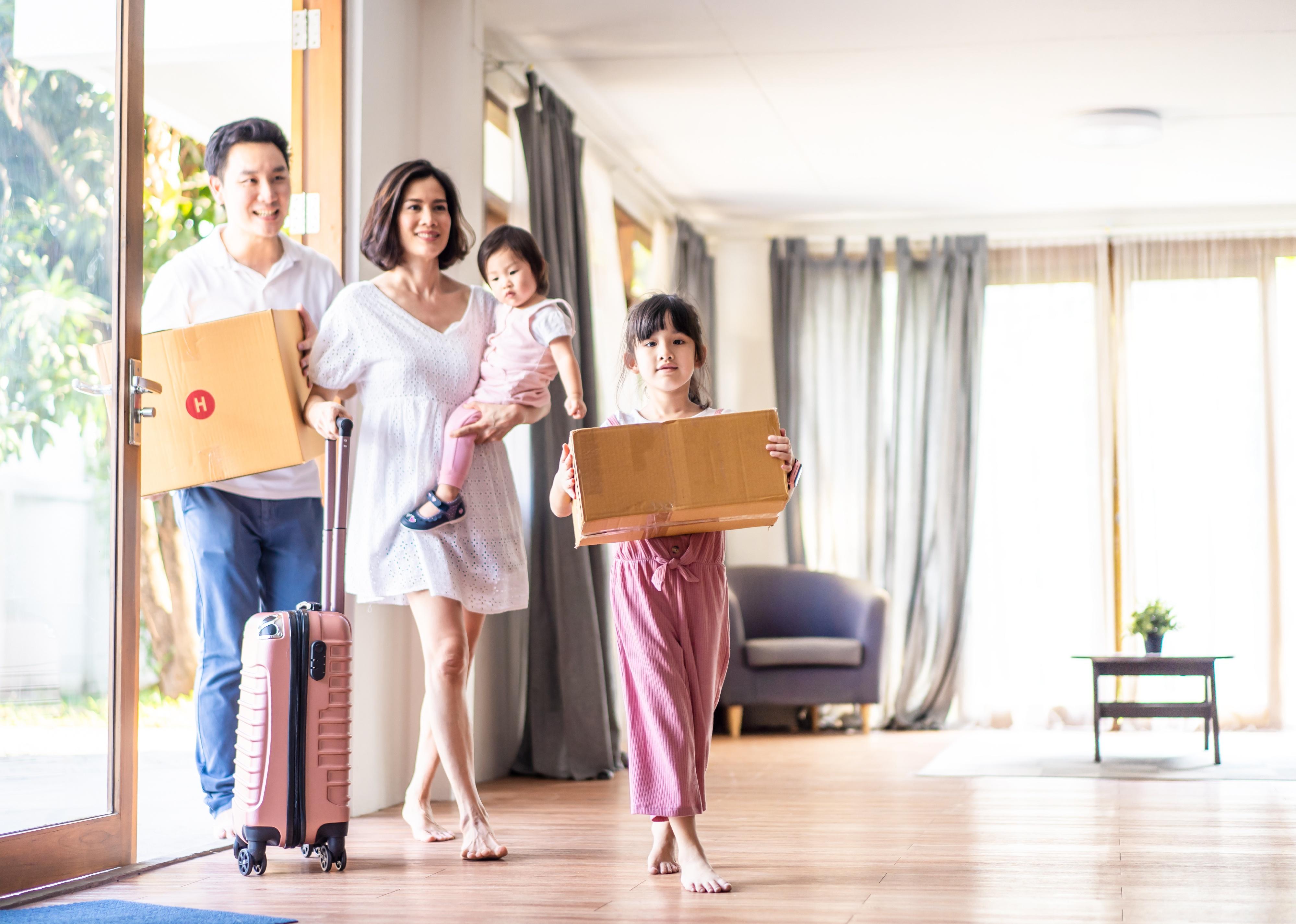 A mother holding a baby girl with suitcase while dad and bigger sister carry boxes into a house.