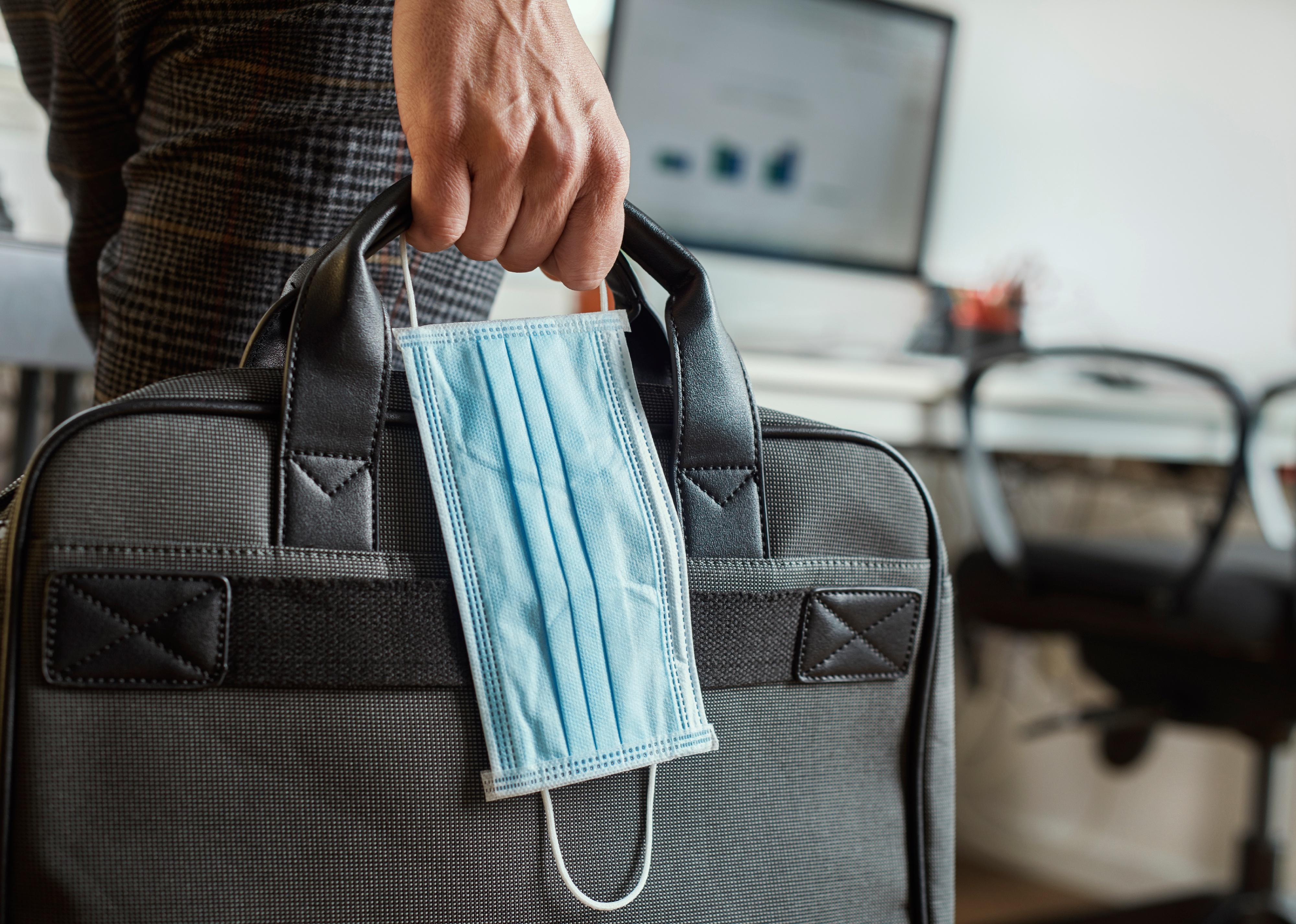 Closeup of a man in an office holding a briefcase and a surgical mask in his hand.