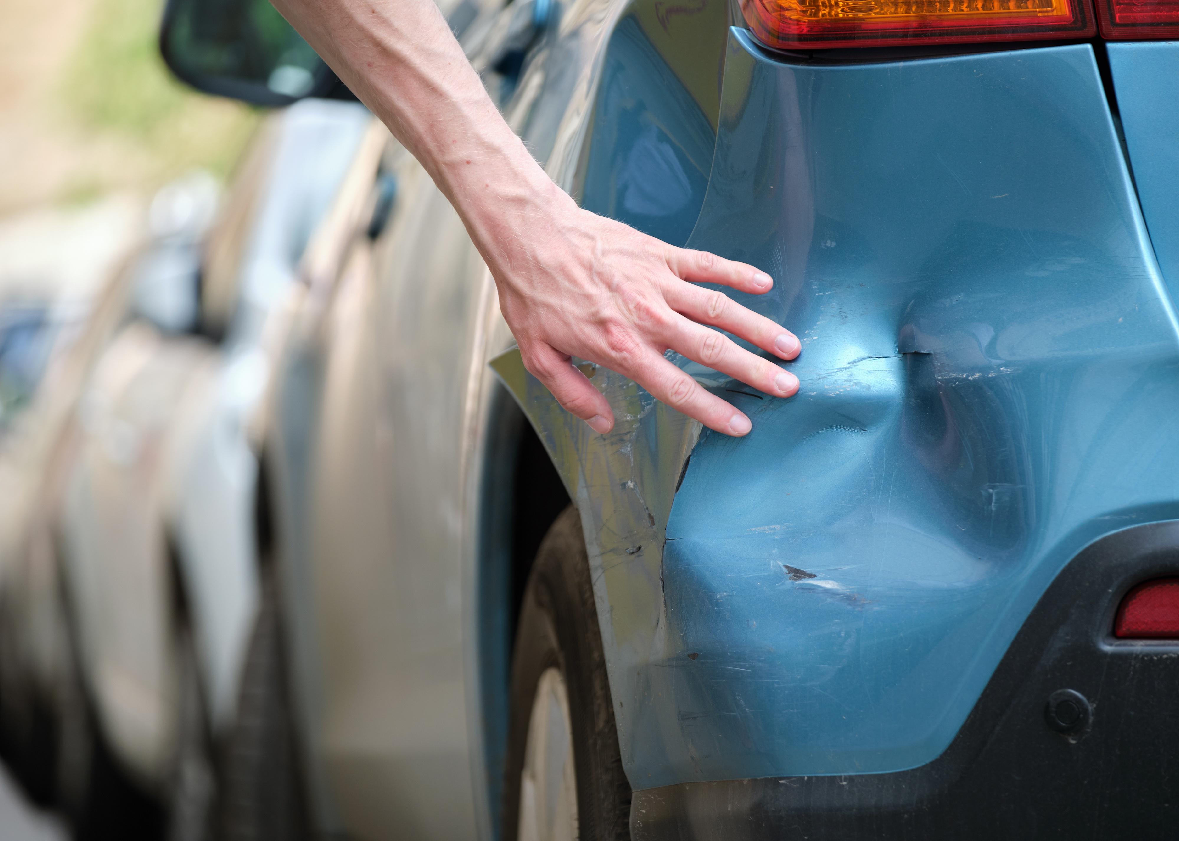 Driver hand examining dented car with damaged fender parked on city street side