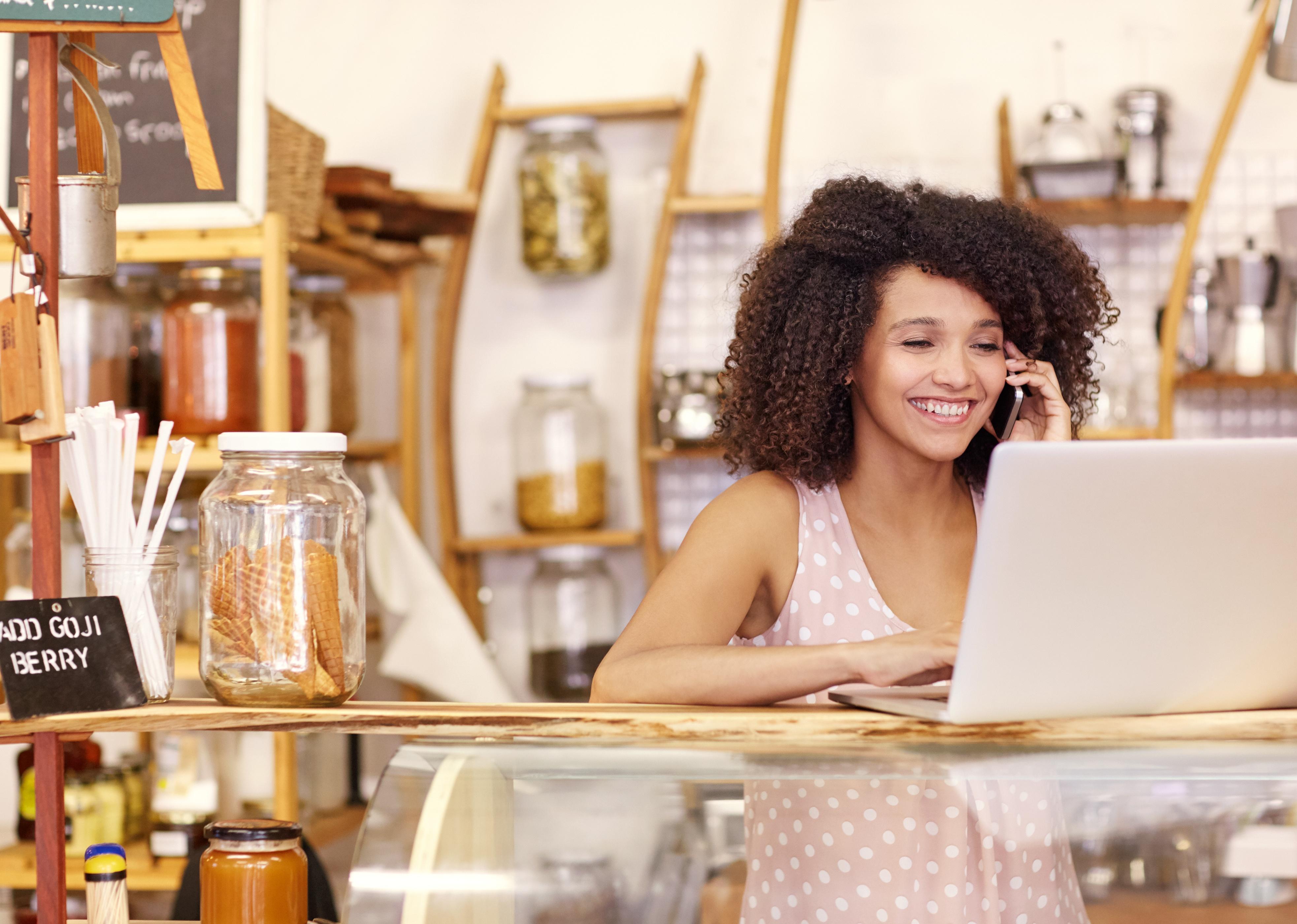 Happy young coffee shop owner talking on phone while typing on their laptop.