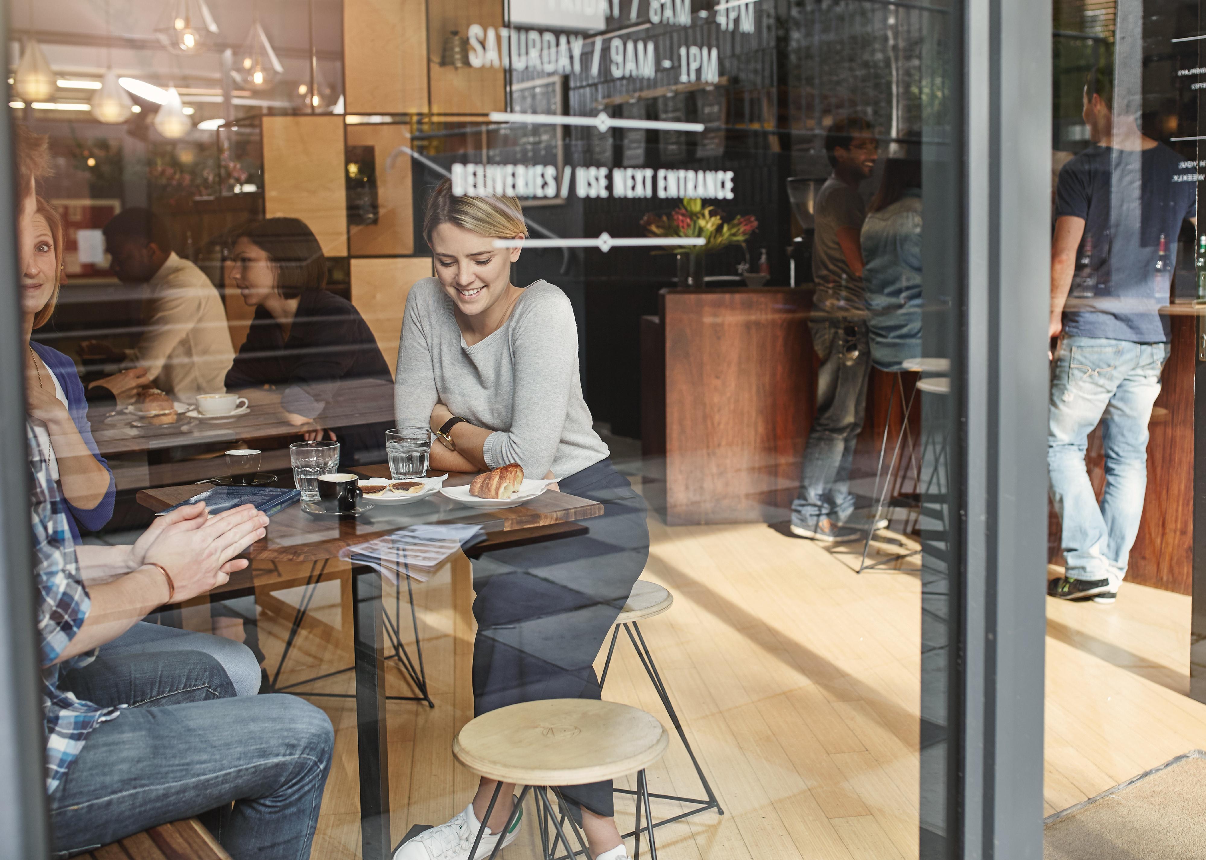 Woman seen through glass window smiling while having coffee with friend.