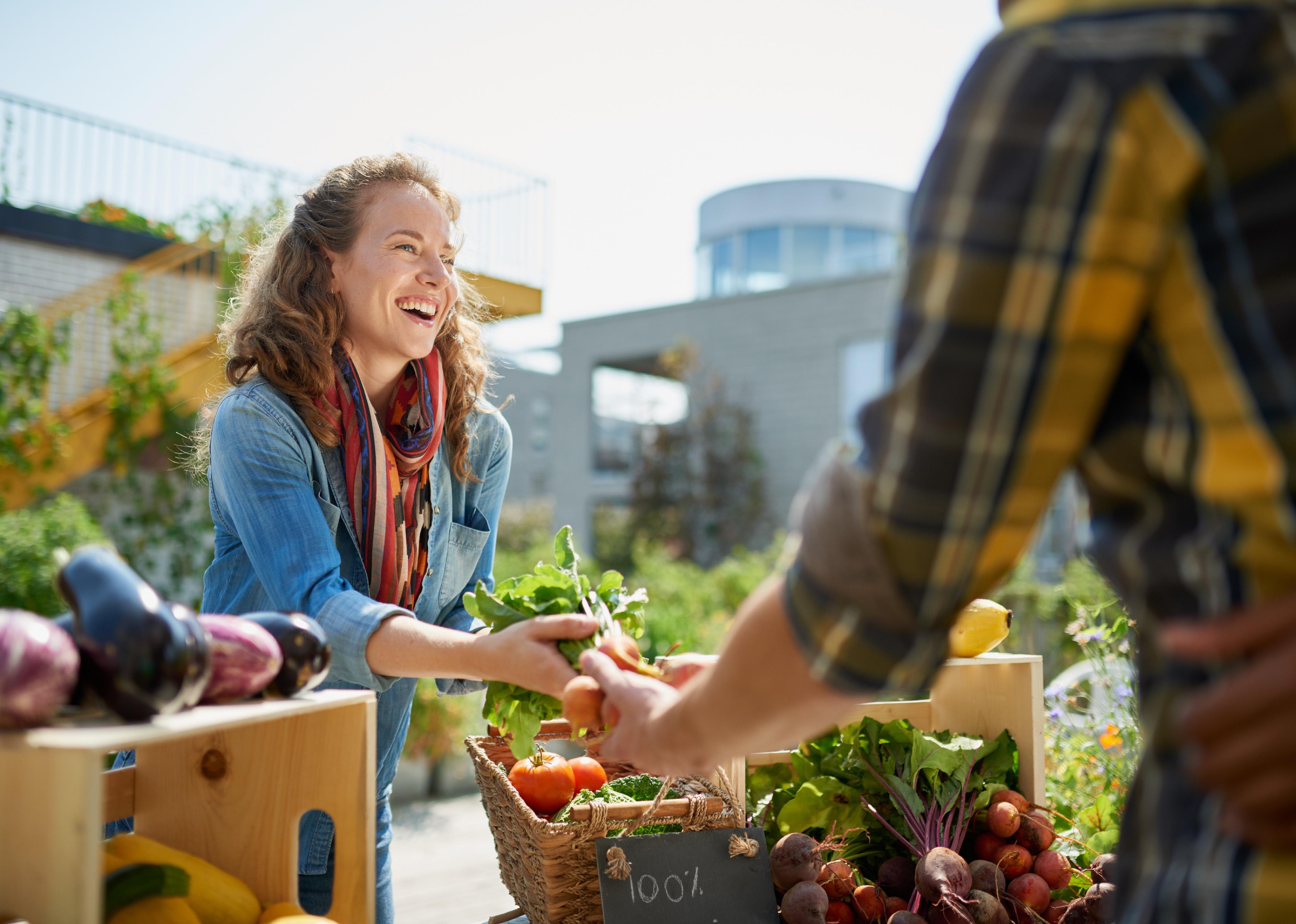 Smiling woman tending a vegetable stall at a farmer's market.