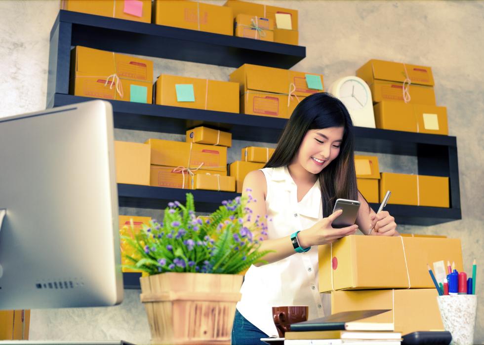 Business owner writing on packages in a modern shipment room with a large computer monitor.