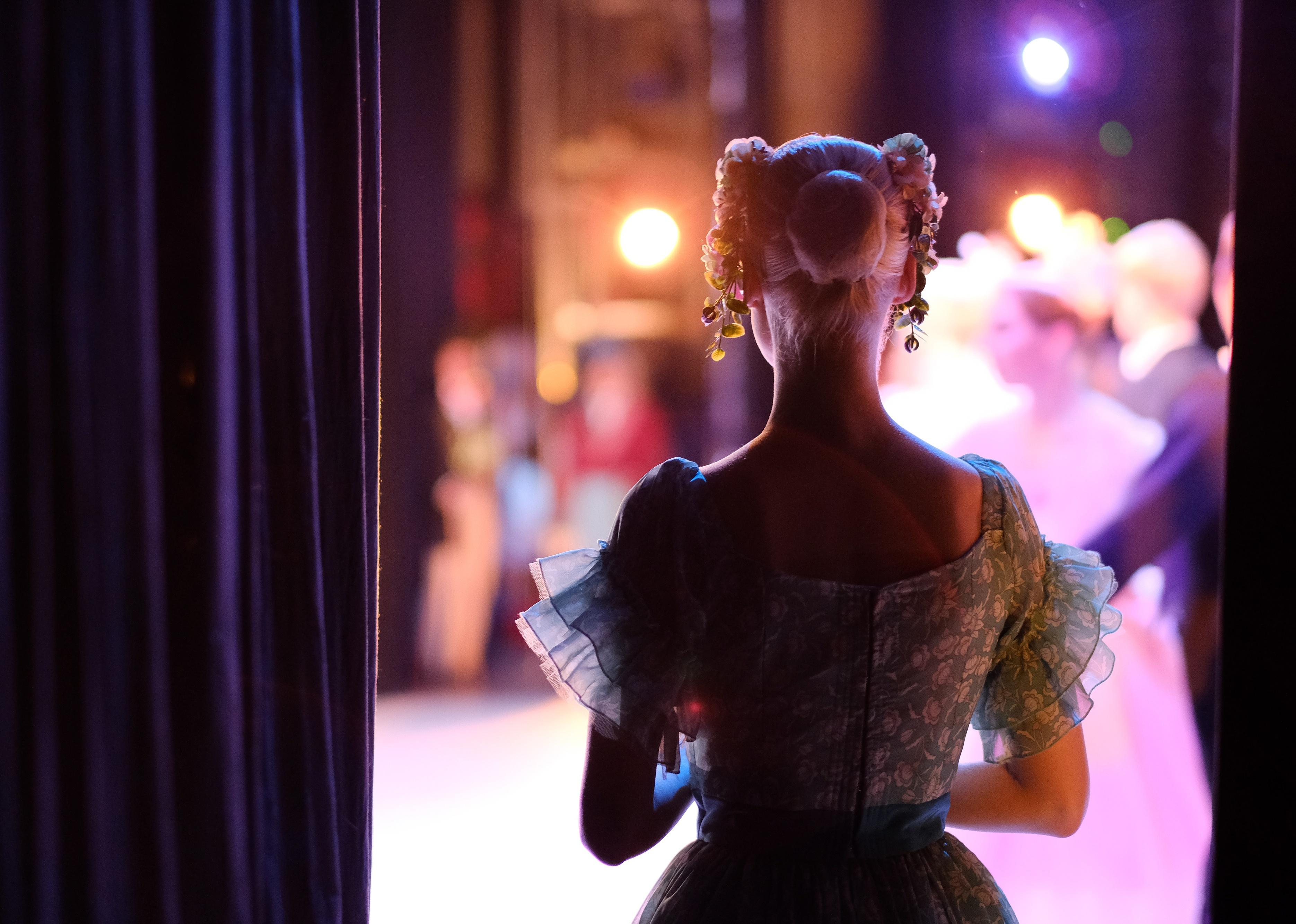 Woman stage actor in costume watches performers through curtain.