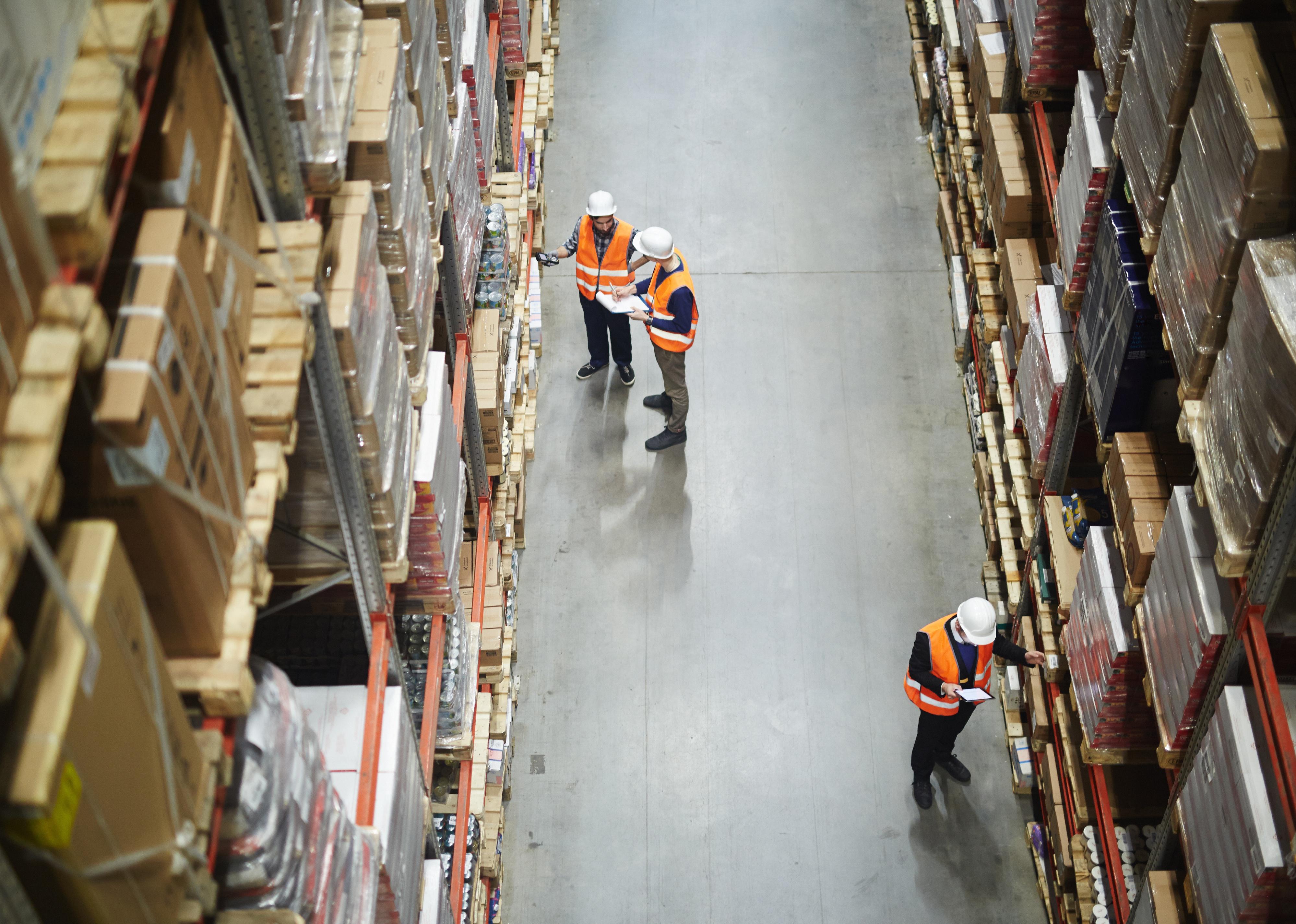 Above view of warehouse workers group in aisle between rows of tall shelves full of packed boxes.